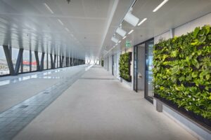 Lichte, moderne overdekte loopbrug aan Joan Muyskenweg met groene plantenwand en grote ramen met stadszicht.